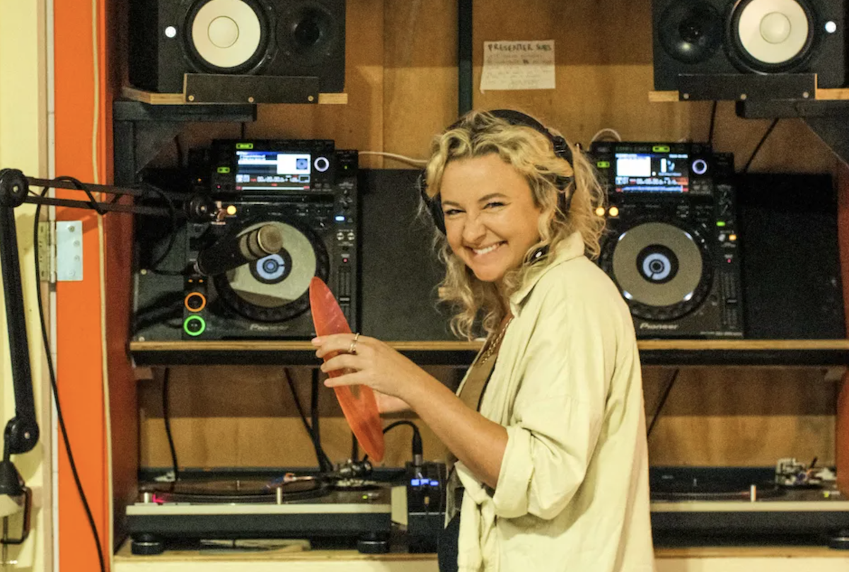 The Porch Sessions founder Sharni Honor smiling with headphones on inside a radio studio booth holding a orange vinyl record next to DJ decks.
